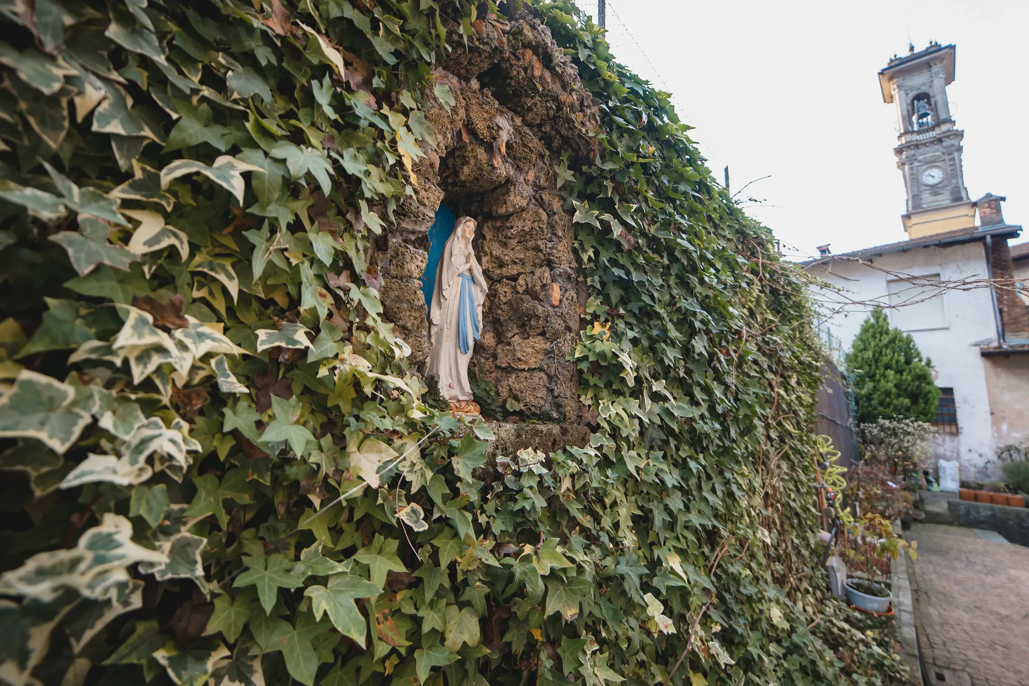 La Madonnina statue set into a stone niche covered in ivy in Porto Ceresio, with the church tower in the background